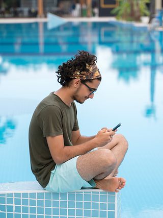 Mann mit einem Smartphone. Die Sonne scheint. Der junge Mann sitzt am Beckenrand eines Pools und surft mobil im Internet. 