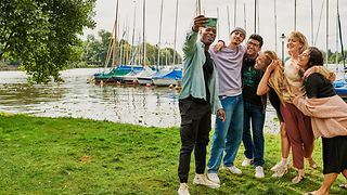 Eine Gruppe junger Menschen macht ein Selfie vor einer ländlichen Kulisse am See. 