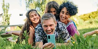 Auf einer Wiese liegende Familie macht ein Selfie mit dem Smartphone.