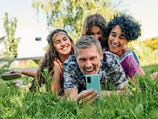 Auf einer Wiese liegende Familie macht ein Selfie mit dem Smartphone.