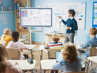 Mehrere Kinder sitzen im Klassenzimmer, die Lehrer steht vor einem digitalen Board.