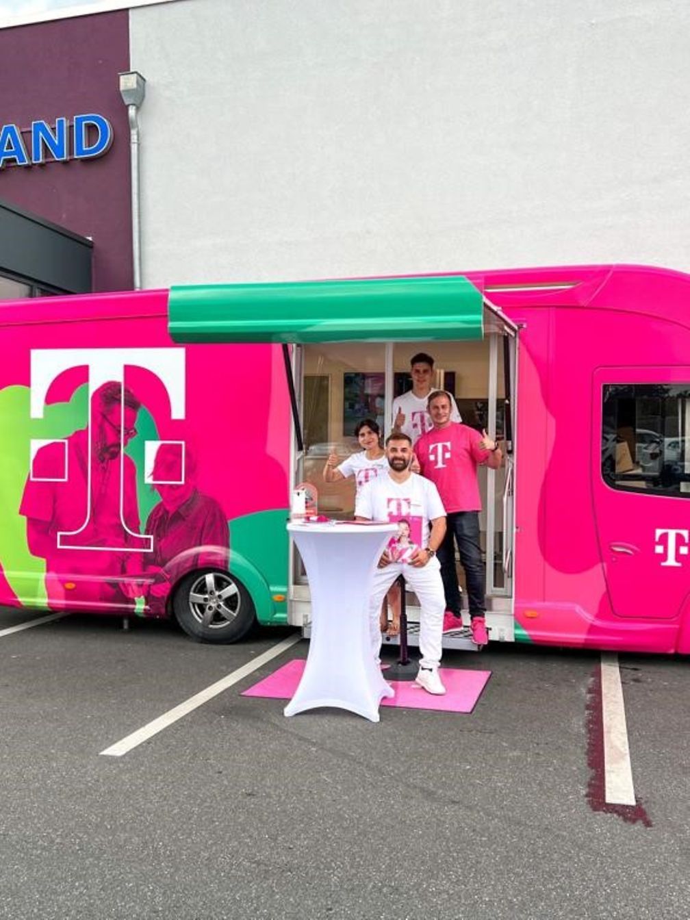 In der geöffneten Tür eines magentafarbenen Trucks steht ein vierköpfiges Beratungsteam und winkt dem Betrachter zu. 