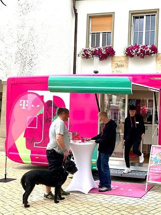 Ein Telekom Mitarbeiter berät einen Kunden mit Hund an einem Stehtisch vor dem Telekom Truck.