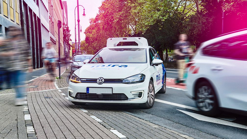 Fahrzeug fährt im Stadtverkehr während Passanten und andere Verkehrsteilnehmende unterwegs sind. 