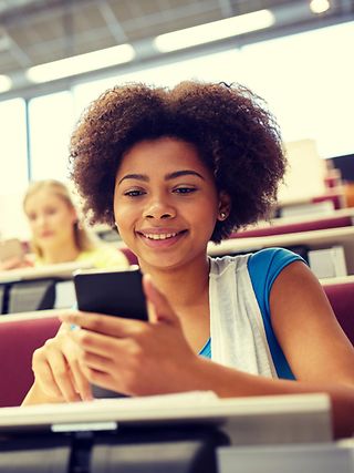 Eine junge Frau sitzt in einem Hörsaal und schaut lächelnd auf ihr Smartphone.