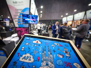 Artist's photo of a tablet with illustrations, including 5G and APIs. Exhibition booths in the background.
