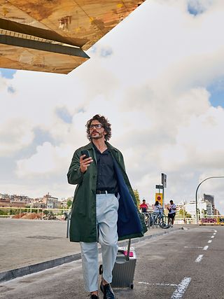 Mann mit Handy in der einen und einem Trolley in der anderen Hand geht eine Straße entlang.