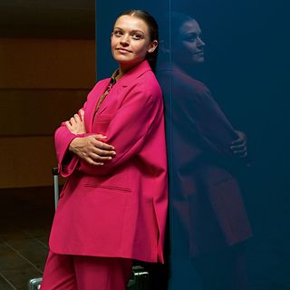Woman in a red jacket leans against a wall, looking into the camera.