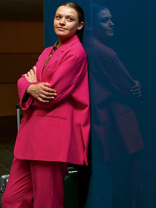 Woman in a red jacket leans against a wall, looking into the camera.