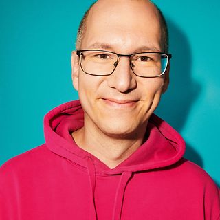 A man in a red hoodie looks into the camera with a friendly smile.