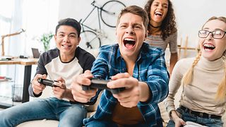 Ein Gruppe junger Menschen spielt euphorisch Videospiele auf dem Sofa sitzend.