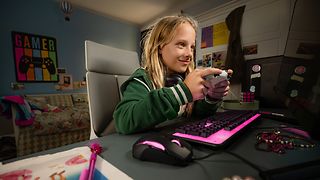 A child sits at a desk and plays computer games. Keyboard and mouse glow in magenta.