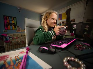A child sits at a desk and plays computer games. Keyboard and mouse glow in magenta.