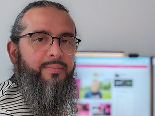  Hasan Kökcü stands in front of his desk with his laptop and two large screens