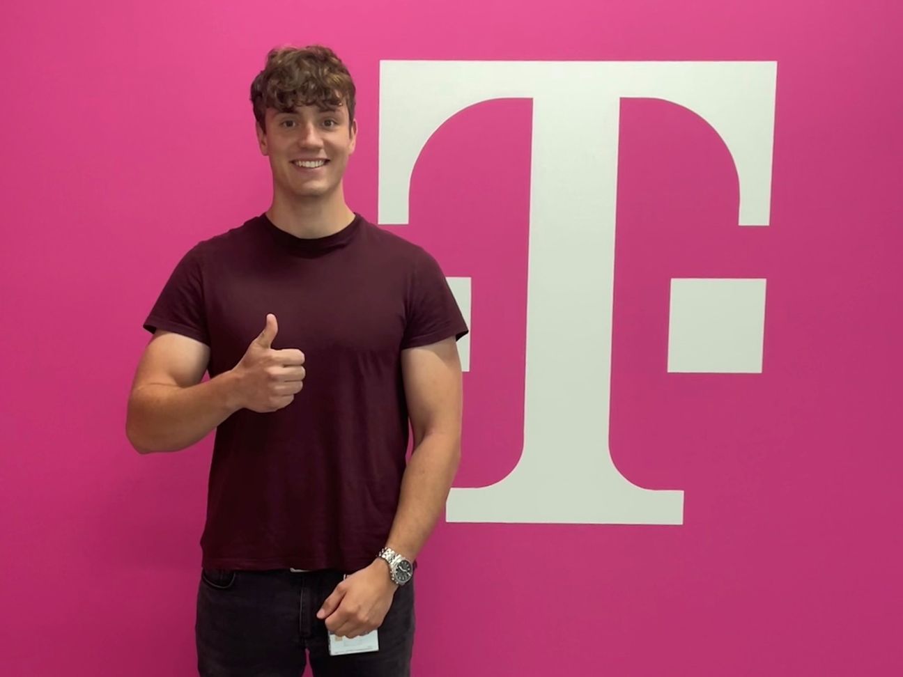 junger Mann zeigt "Daumen hoch" vor einer Wand mit Telekom-Logo.