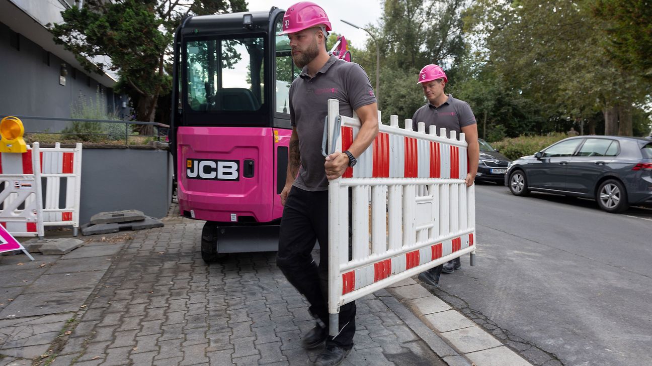 Zwei Männer tragen eine Absprerrung, im Hintergrund steht ein Bagger