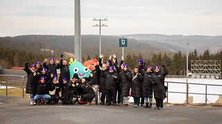 Telekom Mitarbeiterinnen und Mitarbeiter auf einem Berg vor dem Wintersportzentrum in Oberhof, gemeinsam mit dem Maskottchen.