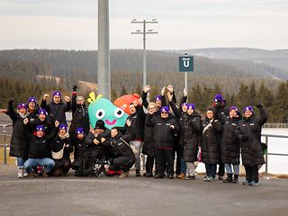 Telekom Mitarbeiterinnen und Mitarbeiter auf einem Berg vor dem Wintersportzentrum in Oberhof, gemeinsam mit dem Maskottchen.