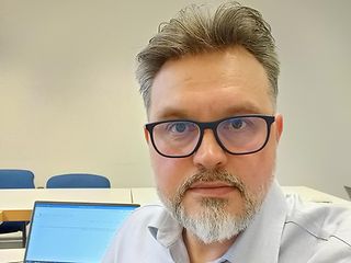 Image of a man sitting at a desk and looking at the camera, with his laptop on the table in the background