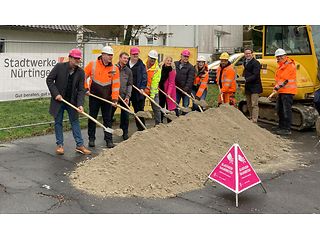 Die Teilnehmer stehen mit Bauhelmen hinter einem großen Sandhaufen und haben symbolisch Schaufeln in der Hand. 