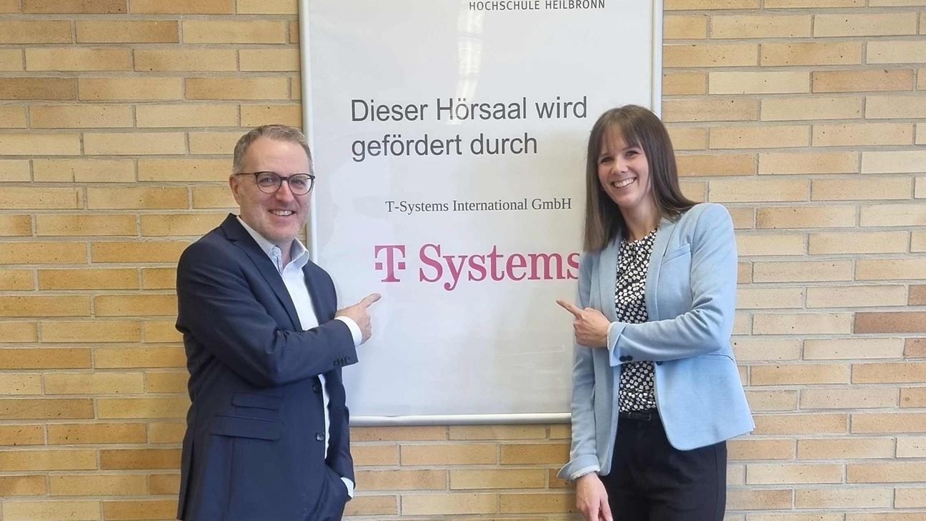 Ein Mann und eine Frau zeigen auf ein Schild mit der Aufschrift „Dieser Hörsaal wird gefördert durch T-Systems“