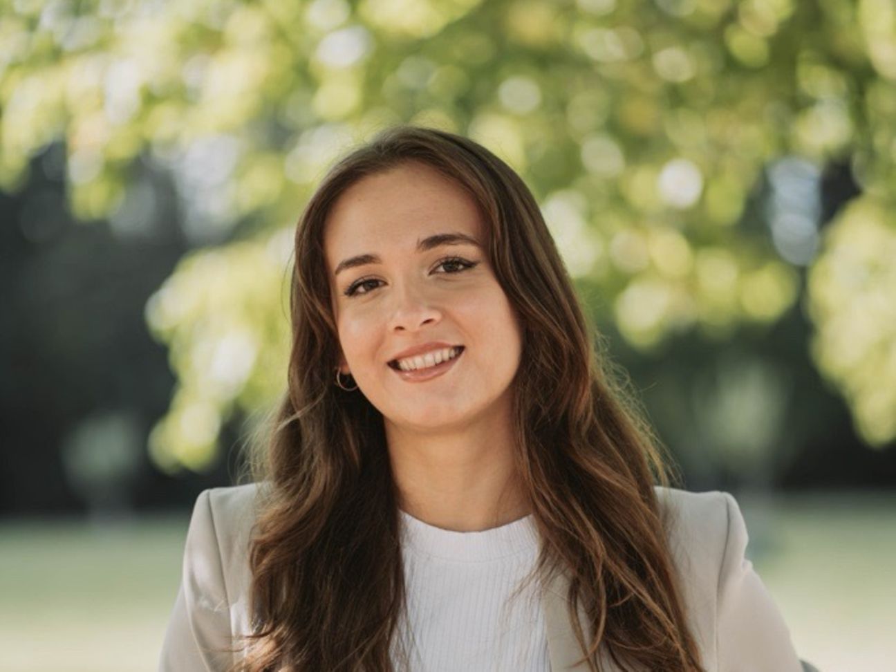 Image of a young woman in nature, looking into the camera, with a background of a meadow and a tree.