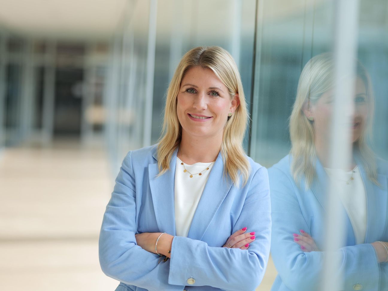 A young woman with long, blond hair, wearing a white shirt and a light blue blazer, smiles into the camera.