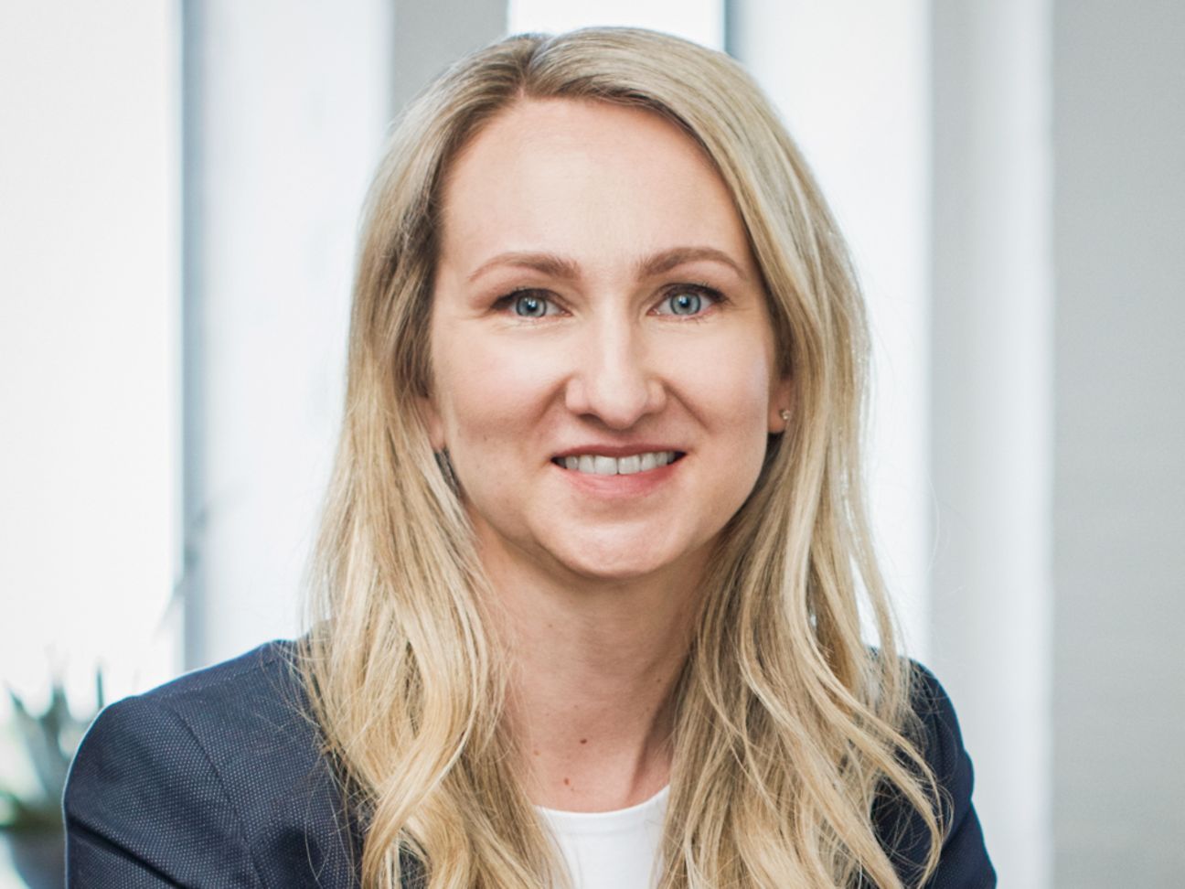 Eine junge Frau mit langen, blonden Haaren trägt einweißes Shirt und einen hellblauen Blazer, lächelt in die Kamera.