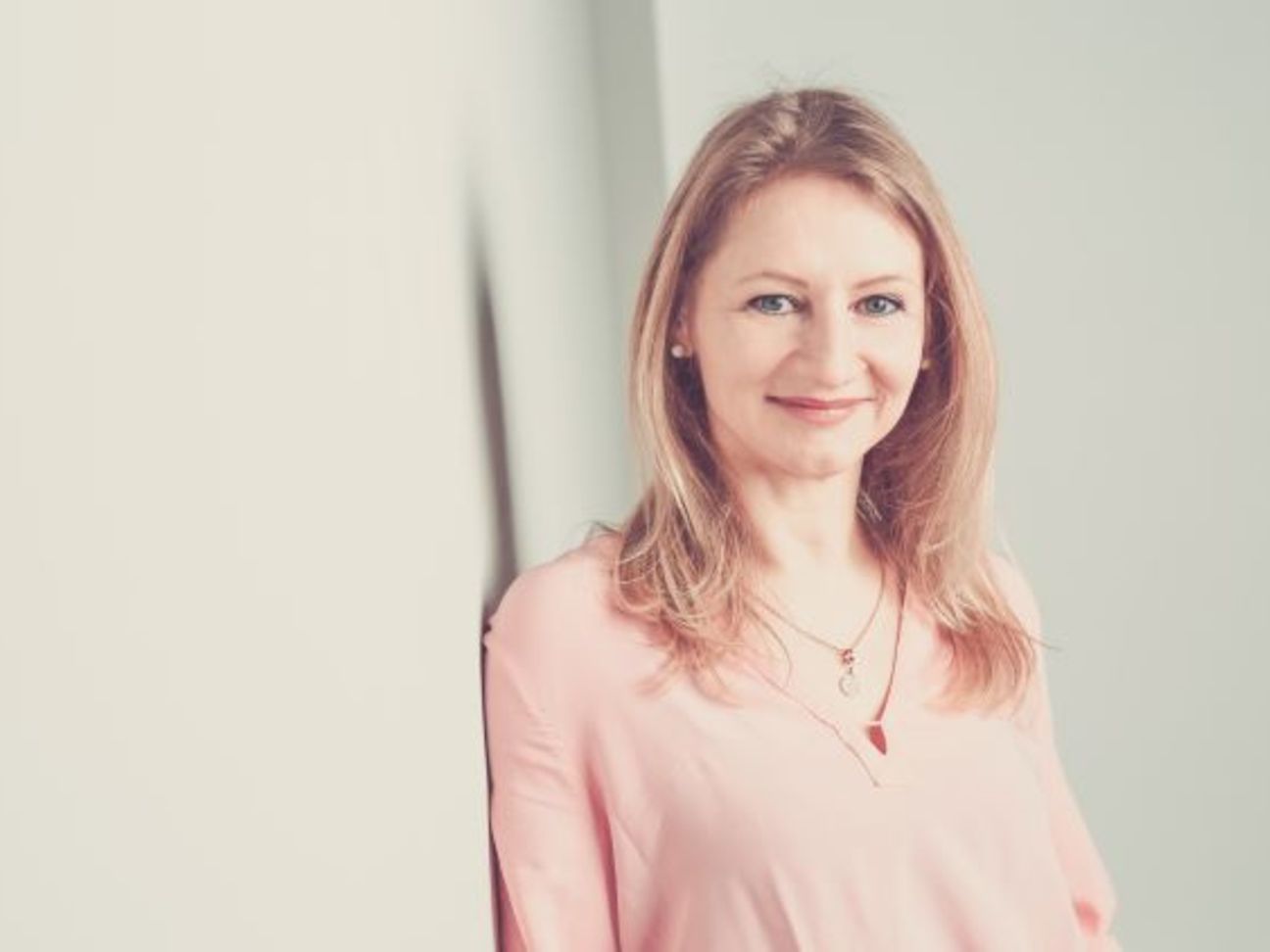 Eine junge Frau lehnt lächelnd an einer hellen Wand und blickt in die Kamera.