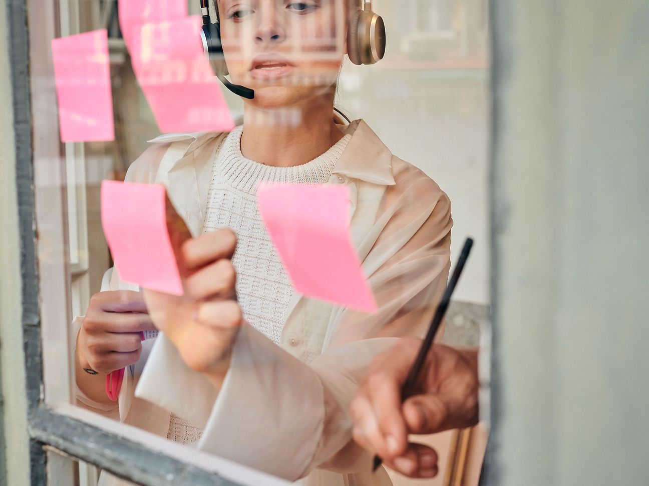 Eine Frau mit Kopfhörern klebt rosa Post-its an eine Scheibe, von rechts ist eine Hand mit einem Stift zu sehen