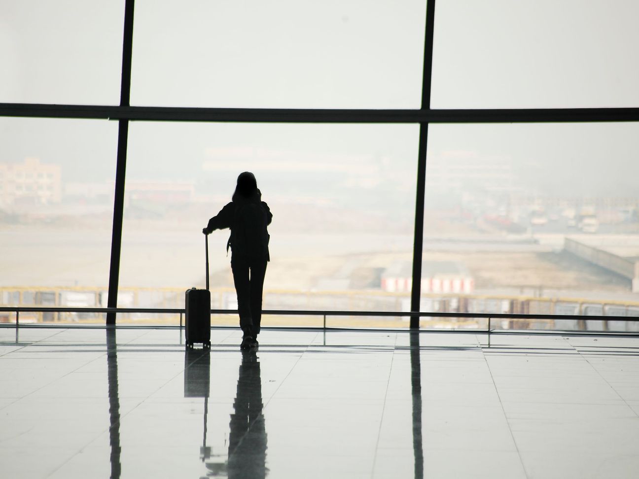A person with a suitcase stands at a large window in the airport and looks at the runway