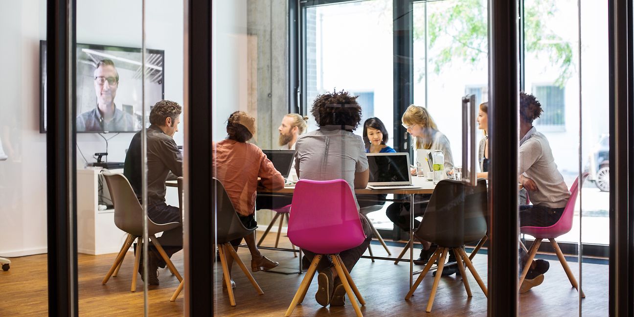 Group of people sitting at a table in a conference room working on laptops, a virtual participant can be seen on a large screen