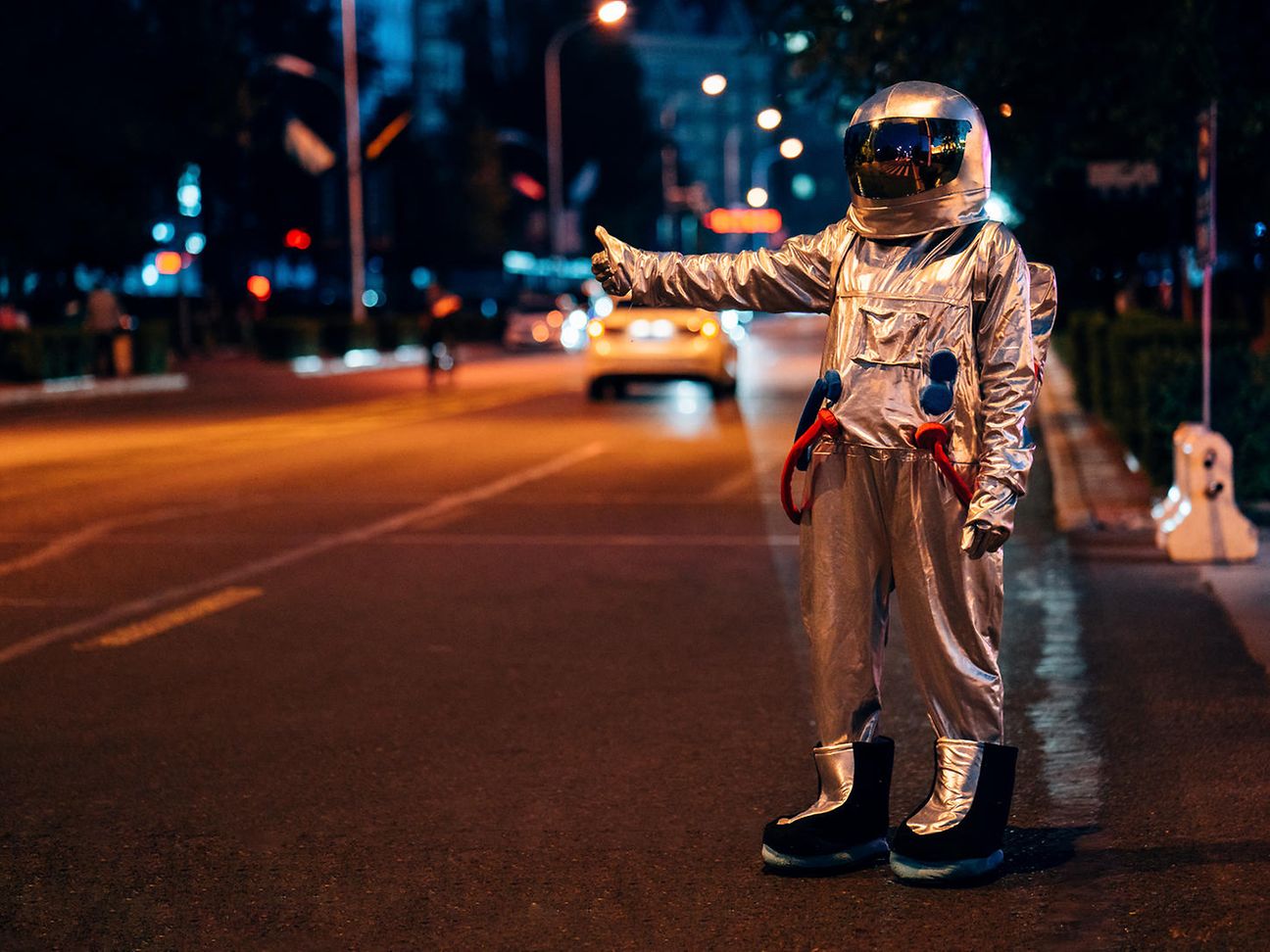 A hitchhiker in a space suit stands in the dark on a road and gives a thumbs up