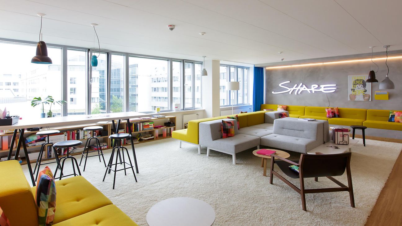 Modern, bright room with yellow sofas, high tables and chairs, and a neon sign stating “SHARE” on the wall