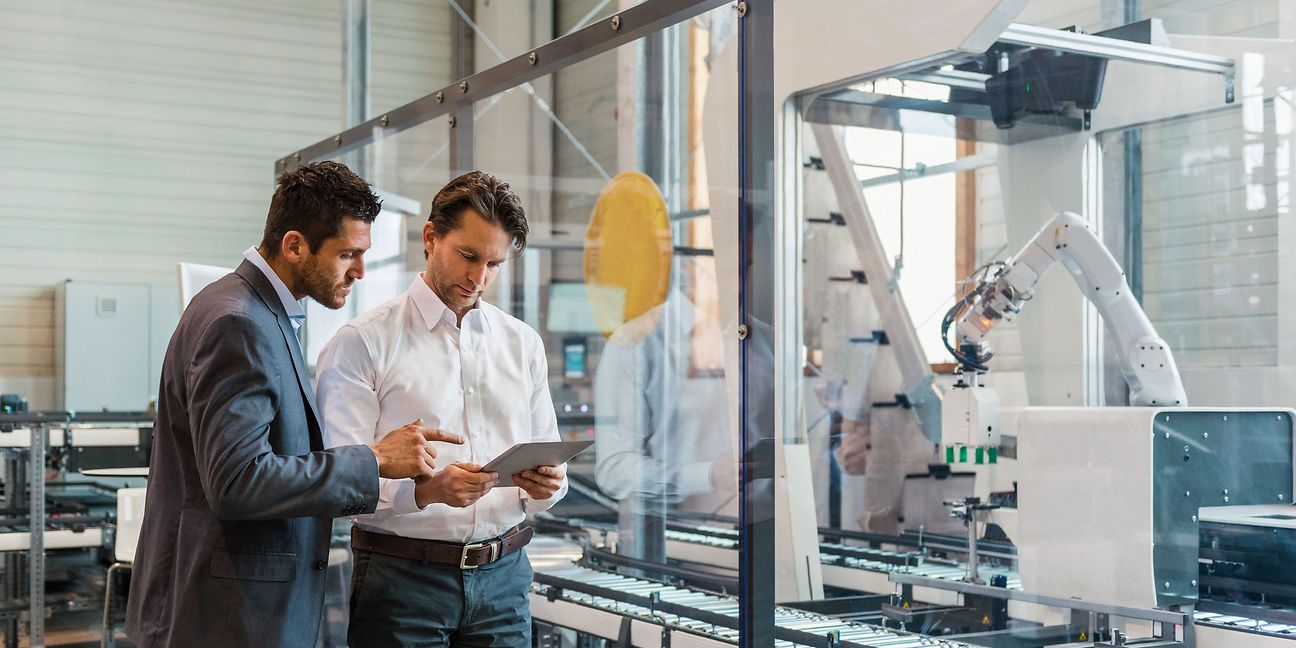 Two men are looking at a tablet together. Machines and a robotic arm can be seen in the background.