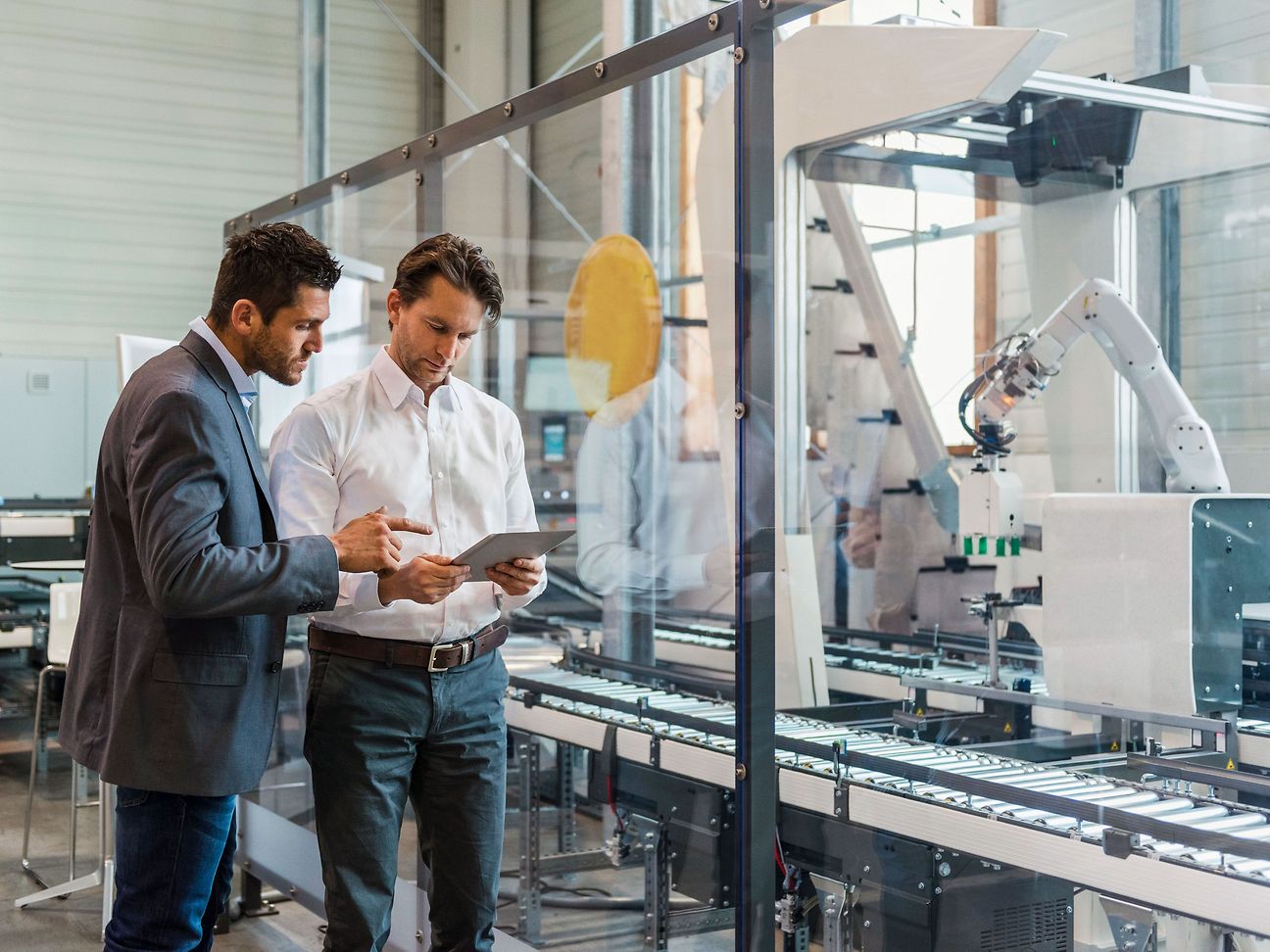 Two men are looking at a tablet together. Machines and a robotic arm can be seen in the background.
