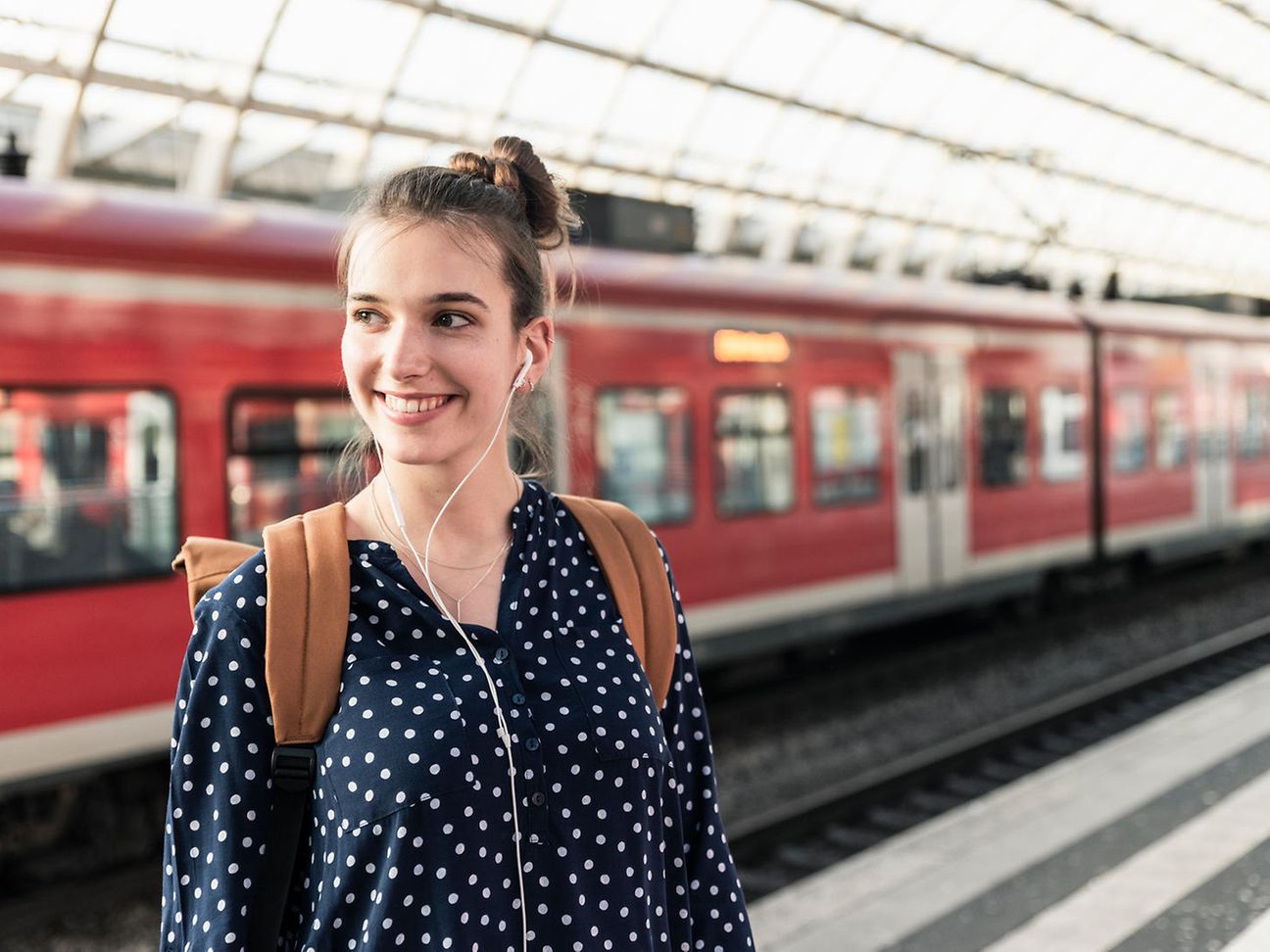 A young woman with a backpack and headphones stands on the platform of a train station.