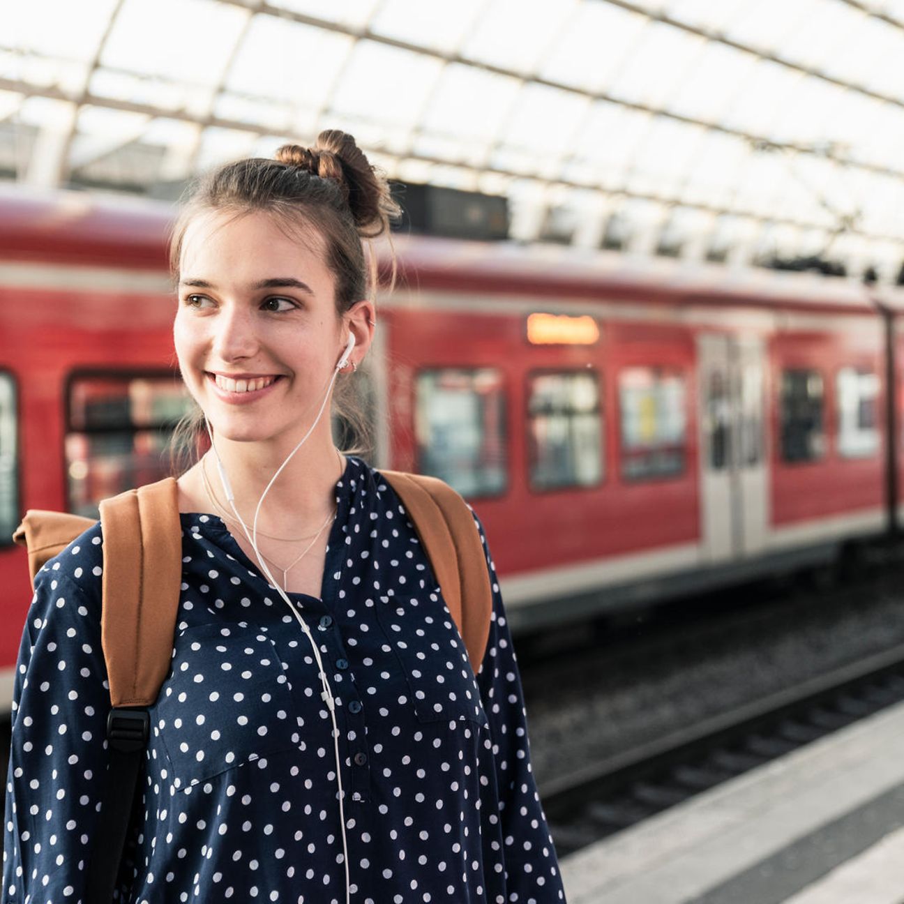 A young woman with a backpack and headphones stands on the platform of a train station.
