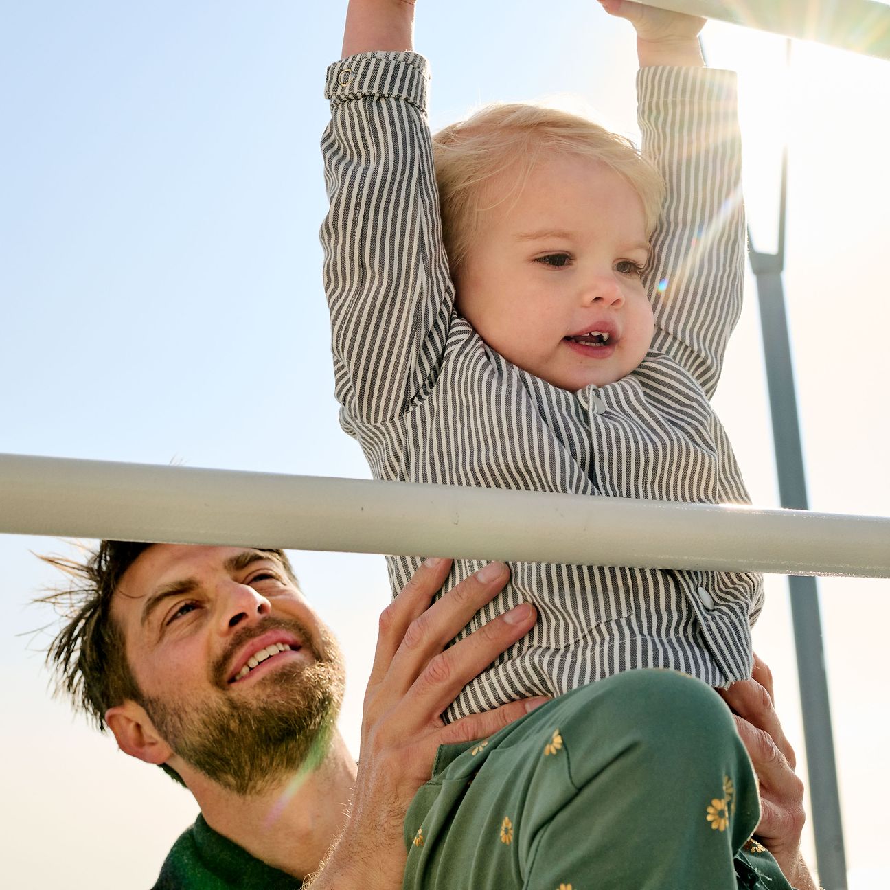 A father is at the playground with his toddler and lifts him onto a climbing frame.