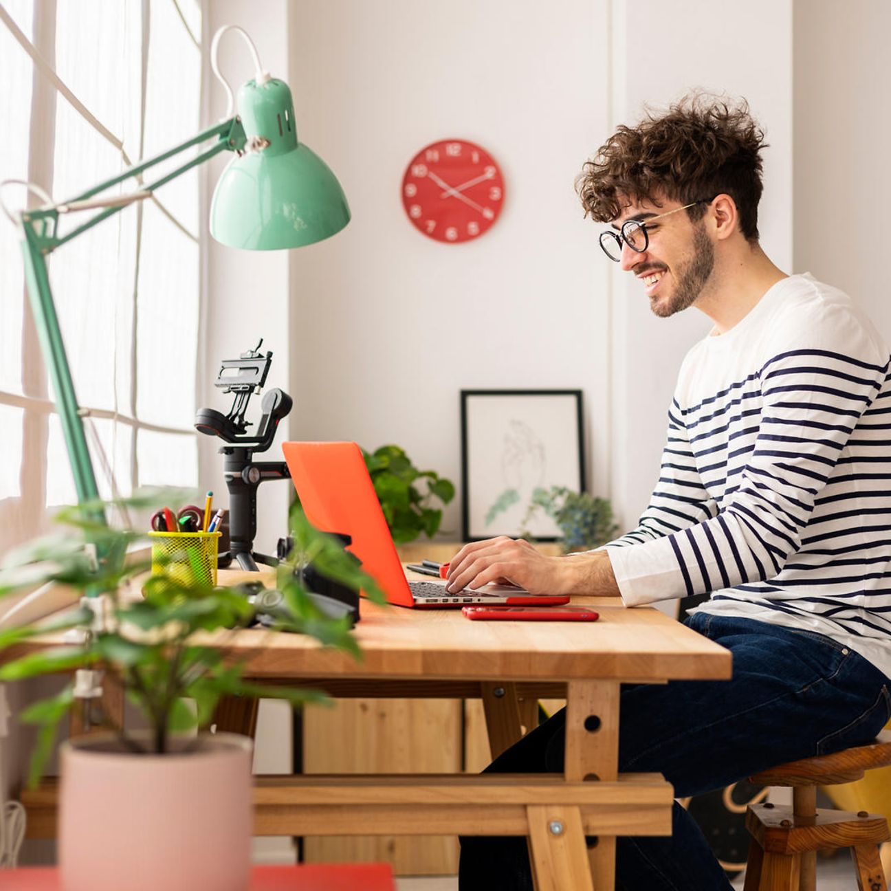 A young man with glasses and a striped sweater works smiling on a laptop in his home