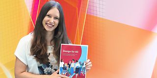 Annika Theiß hält den „Design für Alle Guide“ in der Hand. Sie steht vor einem bunten Design-Hintergrund.