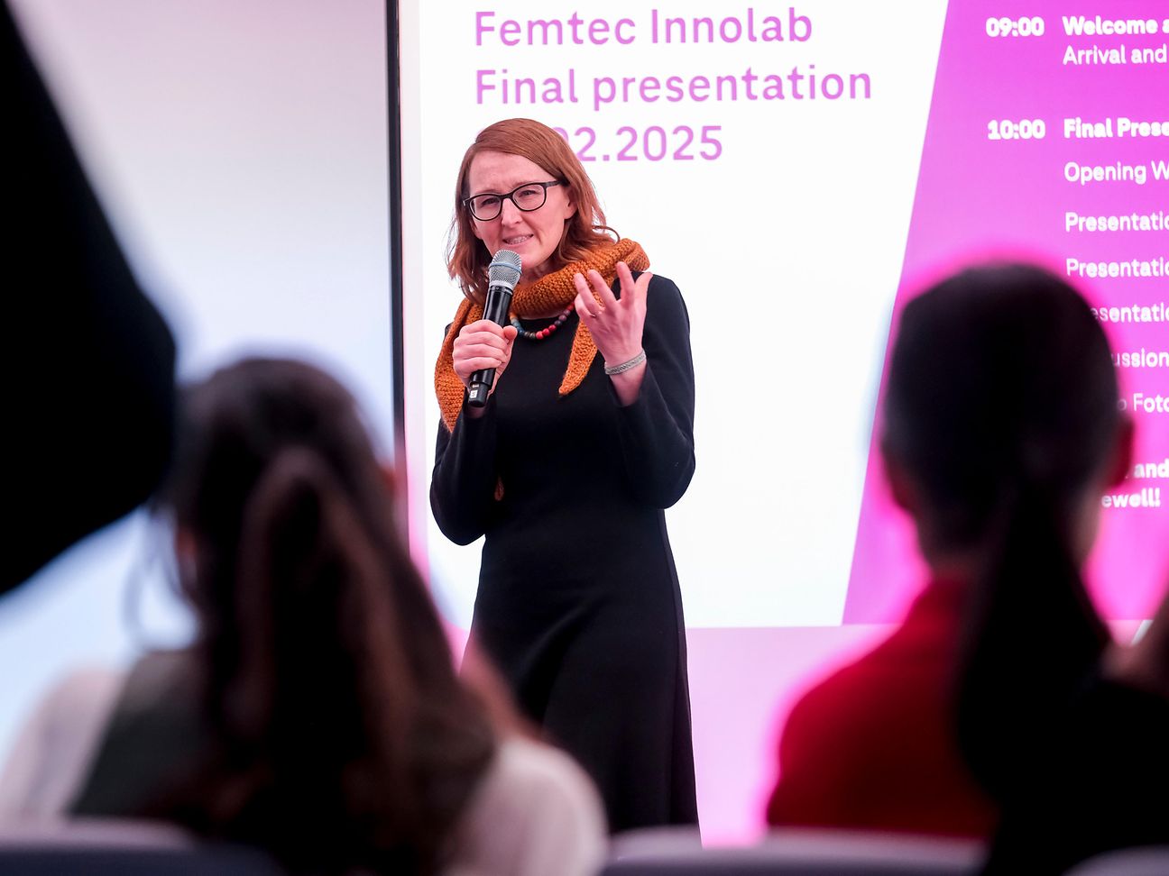 A woman stands in front of a group of women, moderating with a microphone.