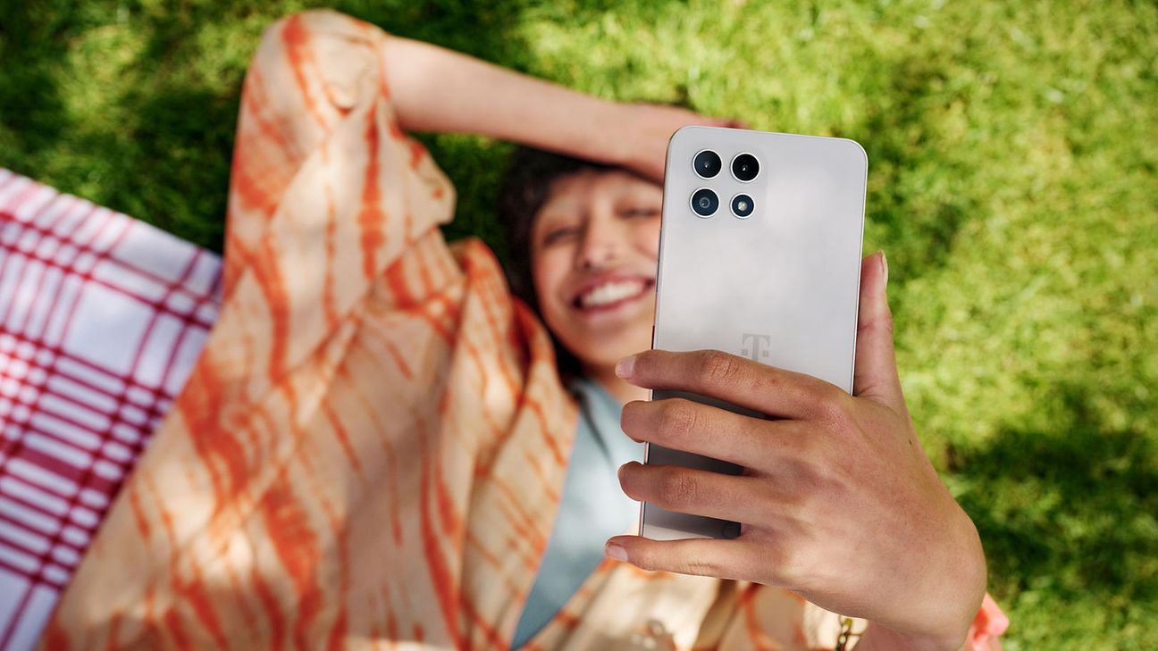 Frau liegt auf der Wiese und lacht und hält ein Handy in die Höhe.