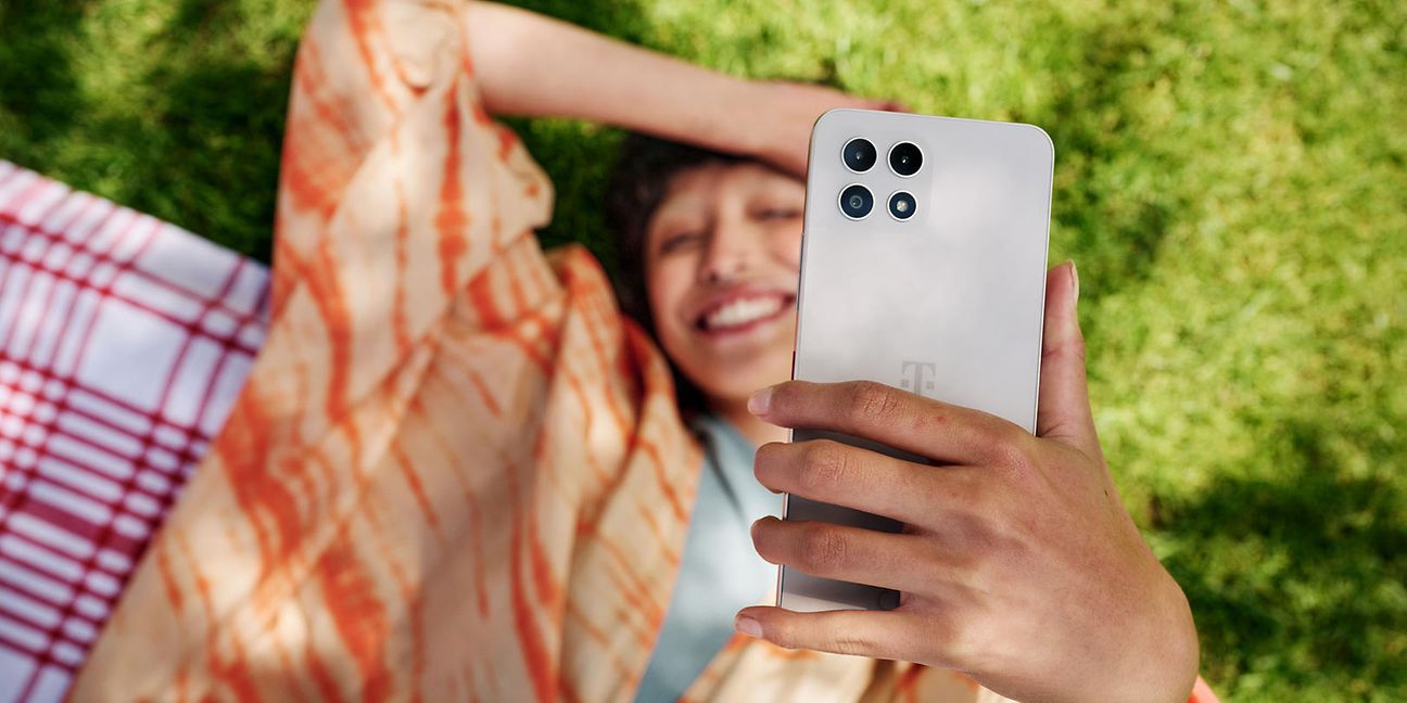 Frau liegt auf der Wiese und lacht und hält ein Handy in die Höhe.