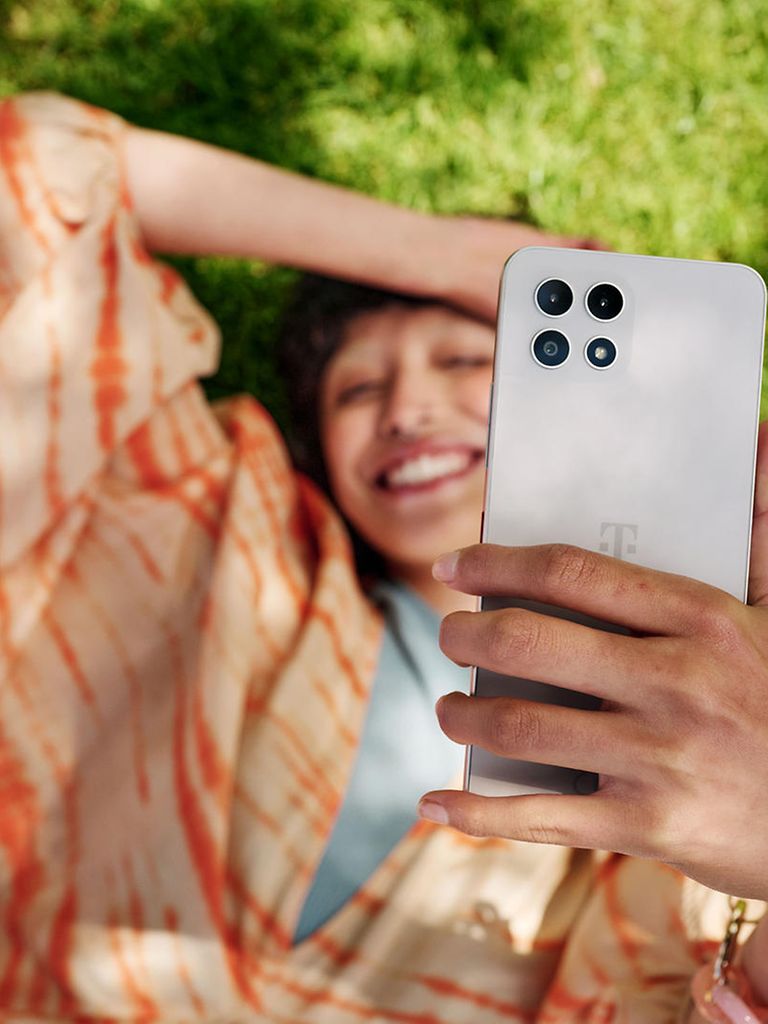Frau liegt auf der Wiese und lacht und hält ein Handy in die Höhe.