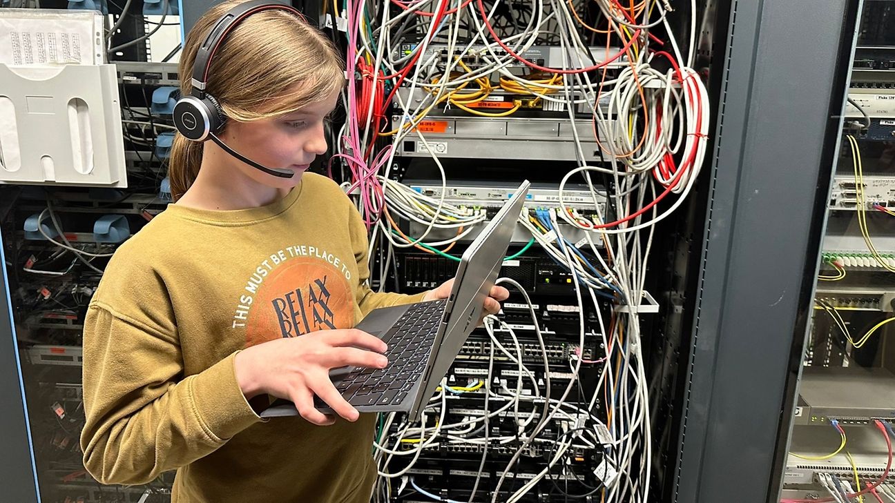 Junges Mädchen hat ein Headset und bedient einen Laptop. Im Hintergrund sieht man viele Kabel in einem Serverraum