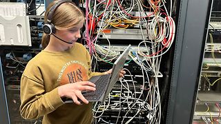 Junges Mädchen hat ein Headset und bedient einen Laptop. Im Hintergrund sieht man viele Kabel in einem Serverraum