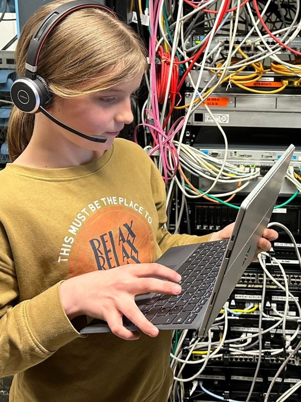 Junges Mädchen hat ein Headset und bedient einen Laptop. Im Hintergrund sieht man viele Kabel in einem Serverraum