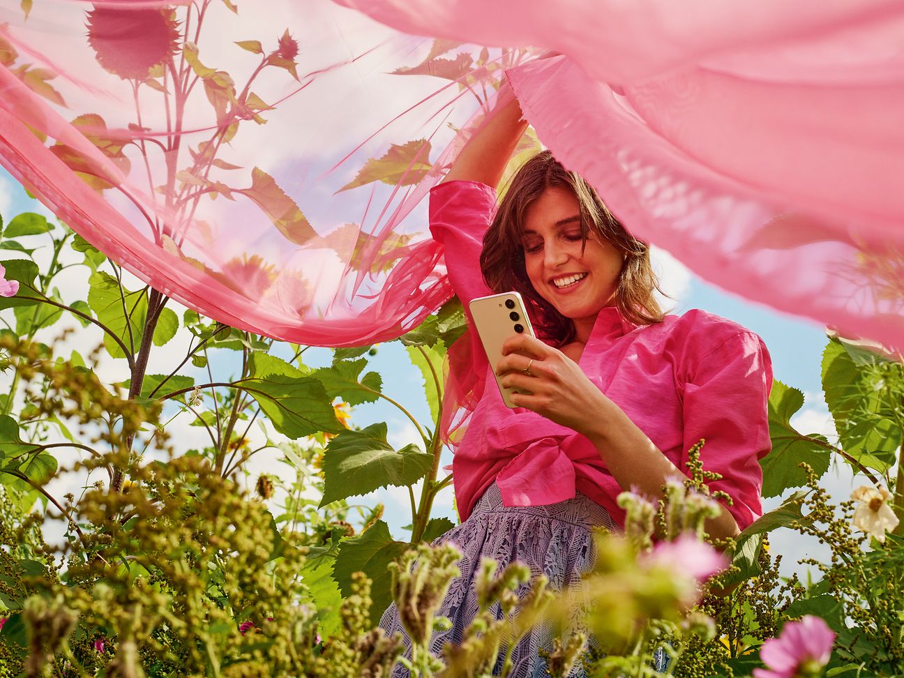 Frau mit Handy und rosafarbenem Schleier steht in einem Blumenfeld.