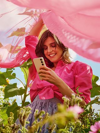 Frau mit Handy und rosafarbenem Schleier steht in einem Blumenfeld.
